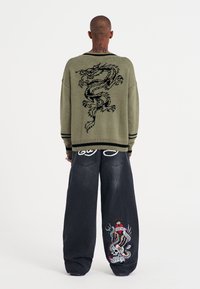 Maglione oversize verde oliva con design di drago nero sul retro. Jeans scuri a gamba larga con dettagli ricamati colorati sulla gamba.