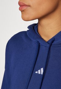Sweatshirt azul marinho com cordões, apresentando um tecido texturizado e um logótipo branco da Adidas na frente. Close-up do decote e dos lábios.