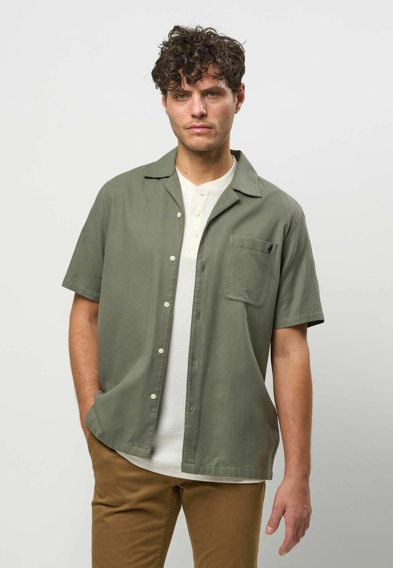 Homme aux cheveux bouclés portant une chemise boutonnée à manches courtes vert olive par-dessus un henley blanc et un pantalon marron, debout devant un fond uni.
