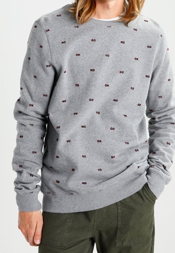 Homme portant un sweat-shirt gris orné de petits textes "OK," "NO," "GO" et "YO", associé à un pantalon vert, mains partiellement dans les poches.