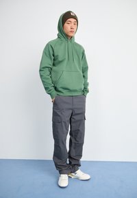 Sudadera verde con un bolsillo frontal, combinada con pantalones cargo grises que tienen bolsillos laterales y zapatillas blancas con acentos amarillos.