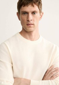 Maglione crema, scollo a girocollo, materiale a maglia leggero, finitura texture, silhouette aderente, indossato con le braccia incrociate su uno sfondo semplice.