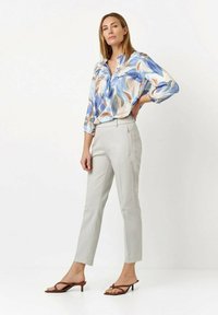 Leichte Bluse mit einem blau-beigen Blättermuster, kombiniert mit hellgrauen, taillierten Hosen und braunen, hochhackigen Sandalen.