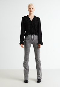 Čierna blúzka s textúrovanými volánmi a širokými manžetami, spárovaná so šedými rozšírenými džínsami. Modelka stojí na neutrálnom pozadí.