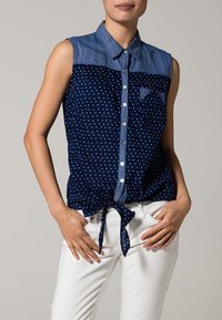 Camicia senza maniche blu navy con colletto azzurro chiaro, pois bianchi, taschino sul petto e orlo annodato. Realizzata in tessuto leggero.