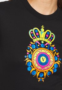 Zwart shirt met een kleurrijk embleem met een kroon en een cirkelvormig ontwerp met de tekst "Versace Jeans Couture" in het midden.