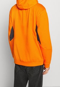 Orange hoodie med svarta sidopaneler, elastisk nederkant och textil med struktur; lös passform med synliga sömdetaljer.