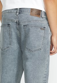 Des jeans en denim bleu clair disposent d'une taille ajustée, de deux poches arrière et d'une étiquette en cuir marron avec des détails de marque sur la ceinture.