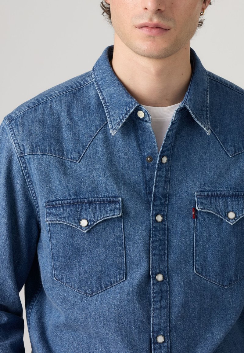 Homme portant une chemise en denim bleu boutonnée avec col pointu et deux poches poitrine par-dessus un t-shirt blanc à col rond.