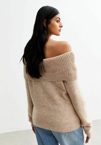 Beżowy sweter bez ramion z ryżowym dekoltem i mankietami. Miękka tekstura, luźny krój, zestawiony z jasnoniebieskimi dżinsami.