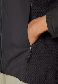 Black jacket featuring a smooth exterior with a textured, zigzag-patterned side. Zippered pocket on the left side with silver hardware.
