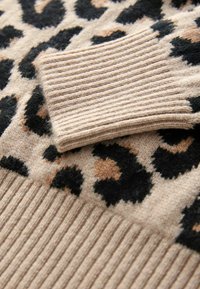 Poignet de manche tricoté beige sur un vêtement en laine à imprimé léopard avec des taches noires et marron et un ourlet côtelé.