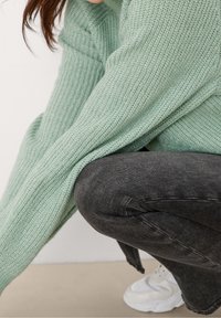 Maglione a maglia verde chiaro con texture e maniche lunghe, abbinato a jeans fitted grigi scuri e sneakers bianche, in posizione accovacciata.