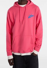 Sudadera rosa hecha de tela suave con un bolsillo frontal. Presenta un logo azul de "Vans". Incluye cordones ajustables y puños acanalados.