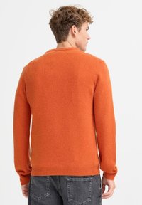 Oransje strikkegenser med lange ermer, rund hals, ribbet mansjetter og kant, myk tekstur, sett bakfra med grå jeans.