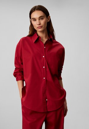 Mujer joven con cabello largo y castaño, vistiendo una camisa roja holgada abotonada y pantalones rojos a juego, de pie con las manos en los bolsillos.