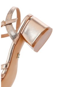 Sandalo in rosa oro metallico con tacco a blocco, cinturino testurizzato e superficie liscia. Dettaglio con fibbia alla caviglia per una vestibilità sicura.