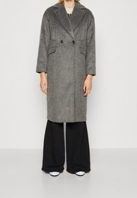 Mujer vestida con un abrigo de lana gris de doble botonadura, sobre pantalones negros de pierna ancha y zapatos blancos, de pie contra un fondo blanco liso.