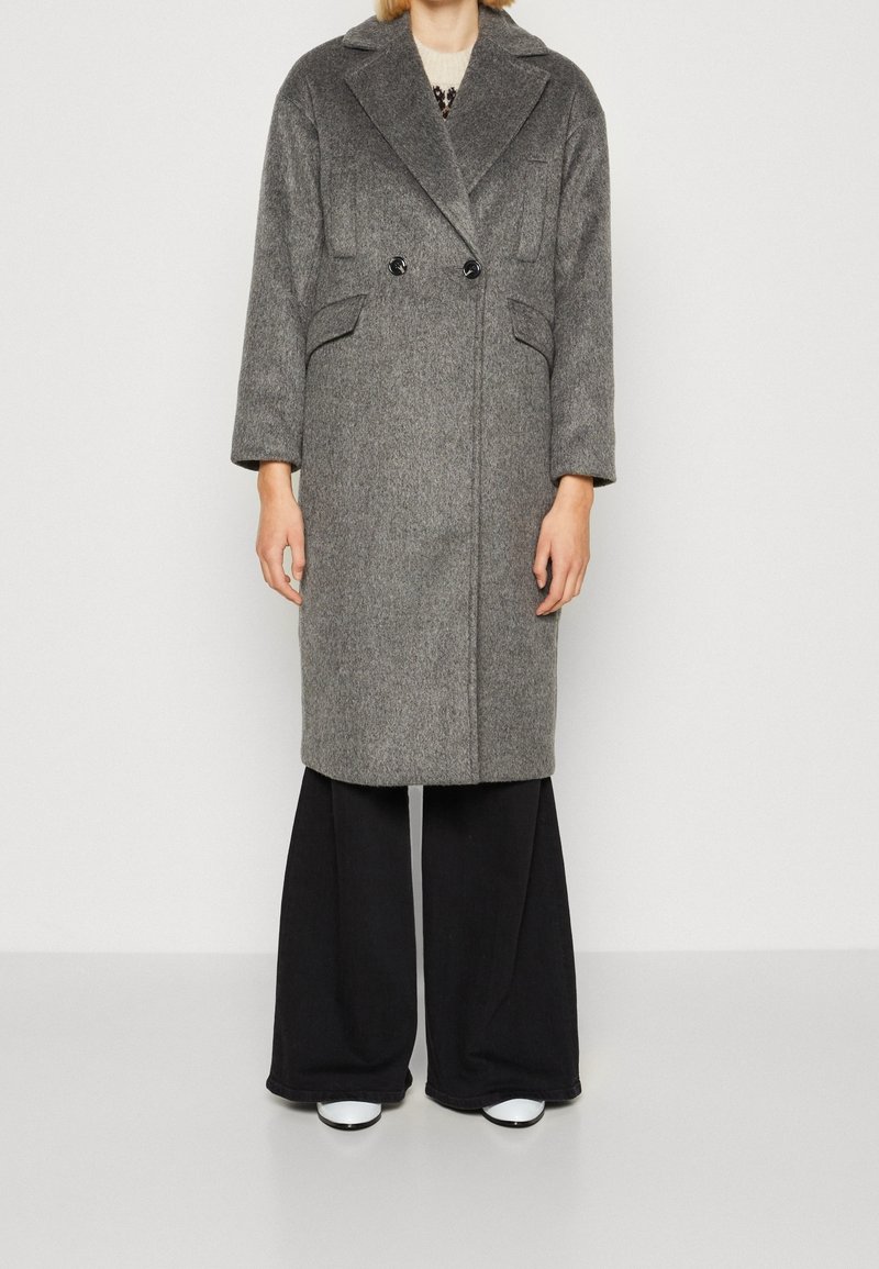 Mujer vestida con un abrigo de lana gris de doble botonadura, sobre pantalones negros de pierna ancha y zapatos blancos, de pie contra un fondo blanco liso.