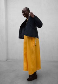 Personne souriante portant une longue robe jaune, une veste noire et des chaussures foncées, debout devant un mur gris uni.