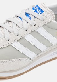 Ljust grå sneaker med vita läderremsor, texturerade detaljer och en blå logotyp. Har en överdel av mocka och nylon, gummisula och platta snören.