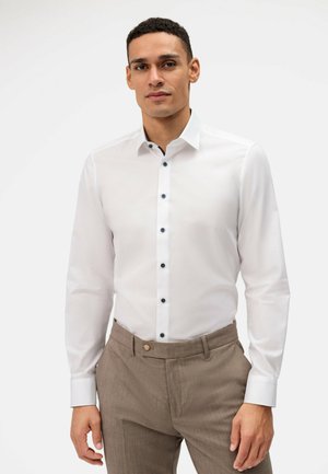 White long-sleeve shirt with a button-down collar and contrasting dark blue buttons. Worn with brown textured trousers.