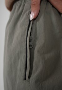 Gros plan sur une main tirant la fermeture éclair d'un tissu vert olive, montrant un matériau lisse et tendu ainsi que de longs ongles manucurés.