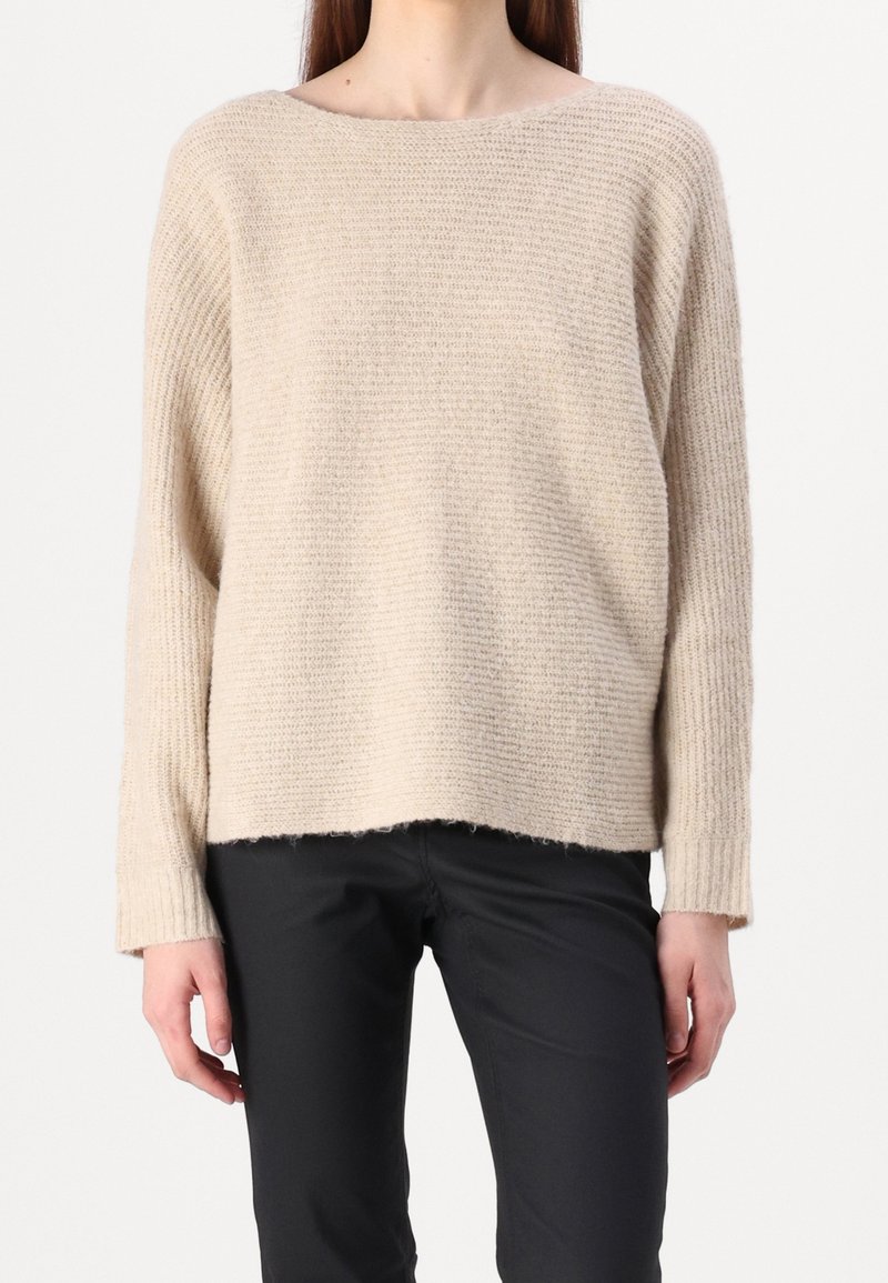 Femme portant un pull tricoté beige ample à manches longues et un pantalon noir, debout devant un fond blanc uni.