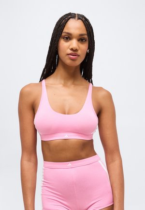 Žena s dlhými pletenými vlasmi, oblečená v ružovom športovom bandeau a zodpovedajúcich vysokým štýlom šortkách s bielymi pruhmi na bočných stranách na neutrálom pozadí.