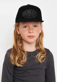Schwarze Baseballmütze mit einem erhabenen, geprägten Batman-Logo auf der Vorderseite. Der Träger hat langes, lockiges Haar und Sommersprossen.