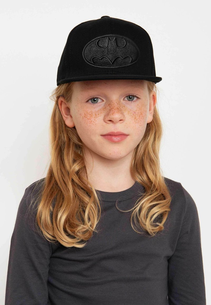 Schwarze Baseballmütze mit einem erhabenen, geprägten Batman-Logo auf der Vorderseite. Der Träger hat langes, lockiges Haar und Sommersprossen.