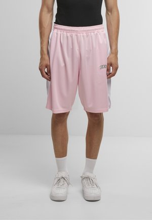 Homme portant un short de basketball en mesh rose avec des panneaux latéraux blancs, des chaussettes blanches et des baskets blanches, debout devant un fond uni clair.