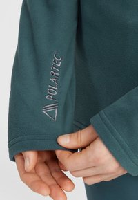 Tyrkysová fleecová bunda s brandingem Polartec na rukávu. Vyznačuje se hladkou texturou a manžetovými rukávy. Detailní záběr na ruku a látku.