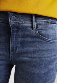 Lähedalt pildistatud sinised teksad esipocketi, vöösahtli ja nupuga, koos kollase kudumisega.