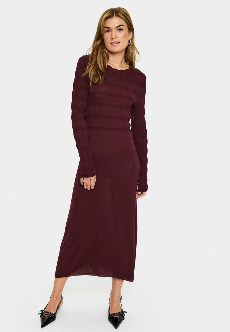 Burgundy knit dress with long, textured sleeves, a fitted bodice, and a flared bottom featuring a central seam detail. Black pointy-toed shoes.