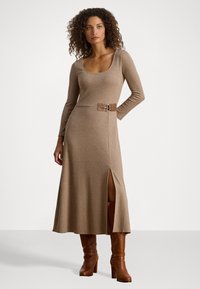 Donna con capelli ricci che indossa un vestito midi beige a maniche lunghe con uno spacco laterale e dettagli in cintura, abbinato a stivali marroni con tacco alti fino al ginocchio.