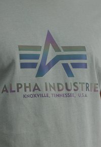 Pilkos medvilnės marškinėliai su daugiaspalviu logotipu, kuriame pavaizduota abstrakti žvaigždė ir horizontalios juostelės. Tekstas skaito "ALPHA INDUSTRIES, Knoxville, Tennessee, JAV."