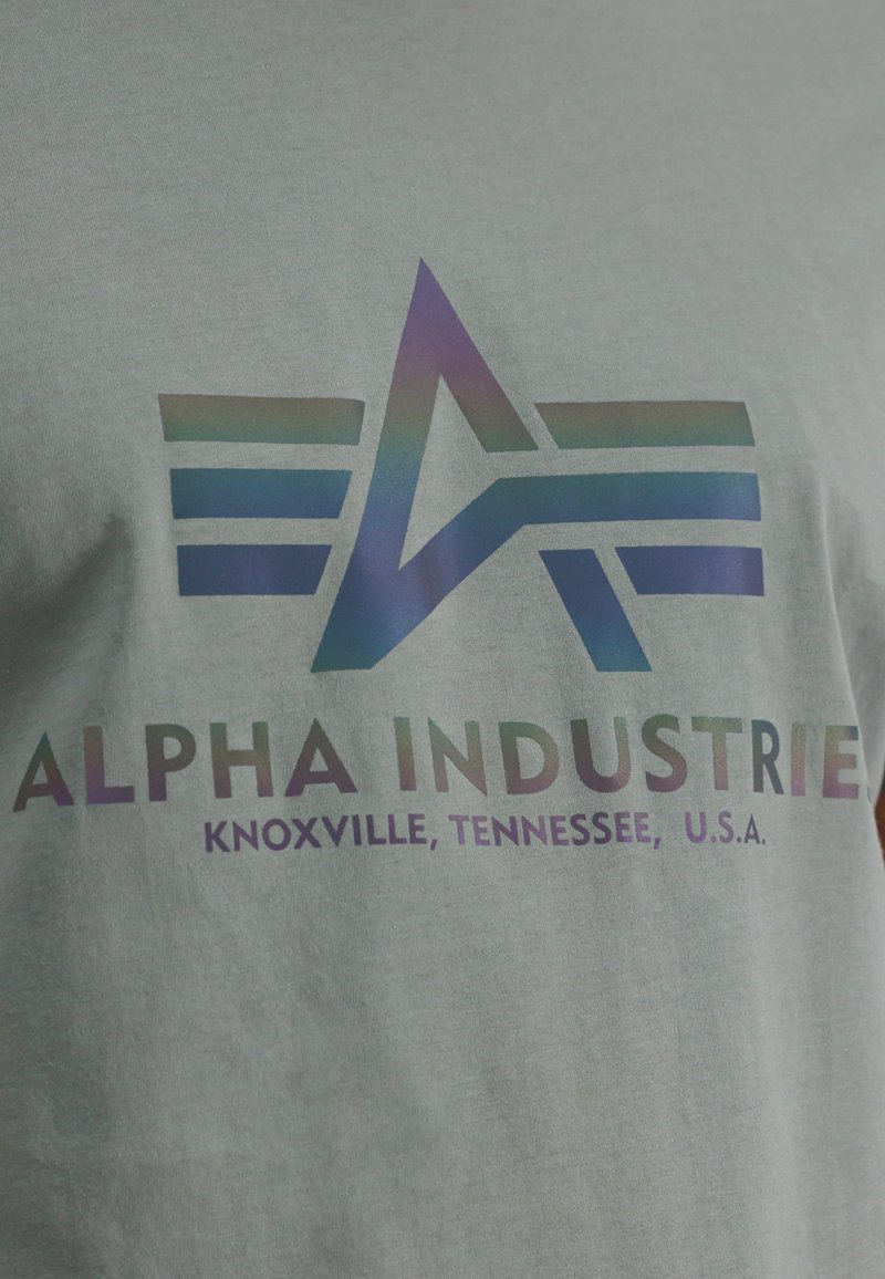 Pilkos medvilnės marškinėliai su daugiaspalviu logotipu, kuriame pavaizduota abstrakti žvaigždė ir horizontalios juostelės. Tekstas skaito "ALPHA INDUSTRIES, Knoxville, Tennessee, JAV."