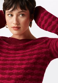 Roter strukturierten Pullover mit einem welligen Muster, weitem Ausschnitt und langen Ärmeln. Modell mit kurzen Haaren und Creolen vor einem neutralen Hintergrund.
