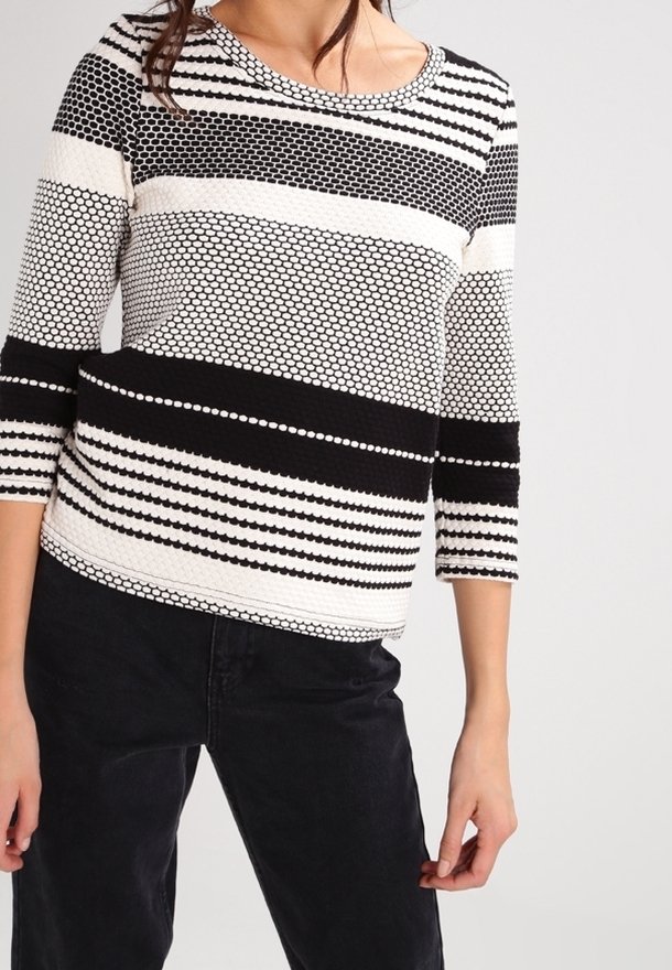 Femme portant un pull en maille à motif noir et blanc avec des manches trois-quarts et un pantalon noir sur un fond uni.
