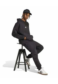 Sudadera negra y pantalones de chándal con puños acanalados, que presentan un pequeño logo blanco. Lleva una gorra negra y zapatillas blancas con detalles en marrón.