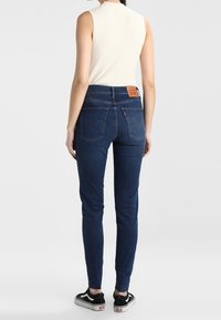 Blaue Skinny-Jeans aus Denim mit hoher Taille, ausgestattet mit Gesäßtaschen und einem hellbraunen Leder-Brandpatch. Kombiniert mit einem hellen ärmellosen Oberteil und schwarzen Sneakers.