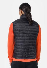 Černá péřová vesta s horizontálním prošíváním, vysokým límcem a lehkým vzhledem, nošená přes oranžové tričko s dlouhým rukávem.