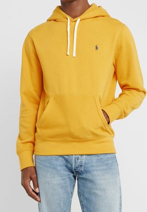 Homme portant un sweat à capuche jaune moutarde avec des cordons blancs et un jean bleu, main dans la poche avant, cadré au niveau du cou et de la taille.