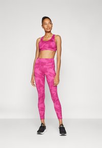 Sostén deportivo de camuflaje rosa y leggings de talle alto hechos de tela elástica, combinados con zapatillas deportivas negras. La modelo está de pie sobre un fondo sencillo.
