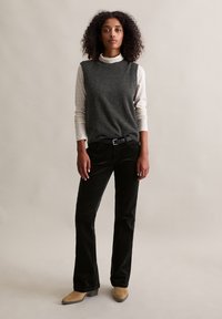 Grau ärmelloser Pullover über einem weißen karierten Hemd, kombiniert mit schwarzen Cordhosen und beigen Knöchelstiefeln. Einfaches, klassisches Design.