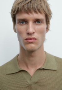 Polo shirt lavorato a maglia verde oliva con colletto, caratterizzato da un motivo testurizzato. Il modello ha i capelli corti castano chiaro e occhi blu chiari.