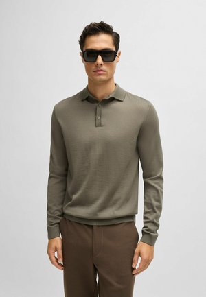 Polo shirt verde oliva a maniche lunghe con texture a costine, colletto con tre bottoni e polsini aderenti, abbinato a pantaloni marroni. Occhiali da sole neri.
