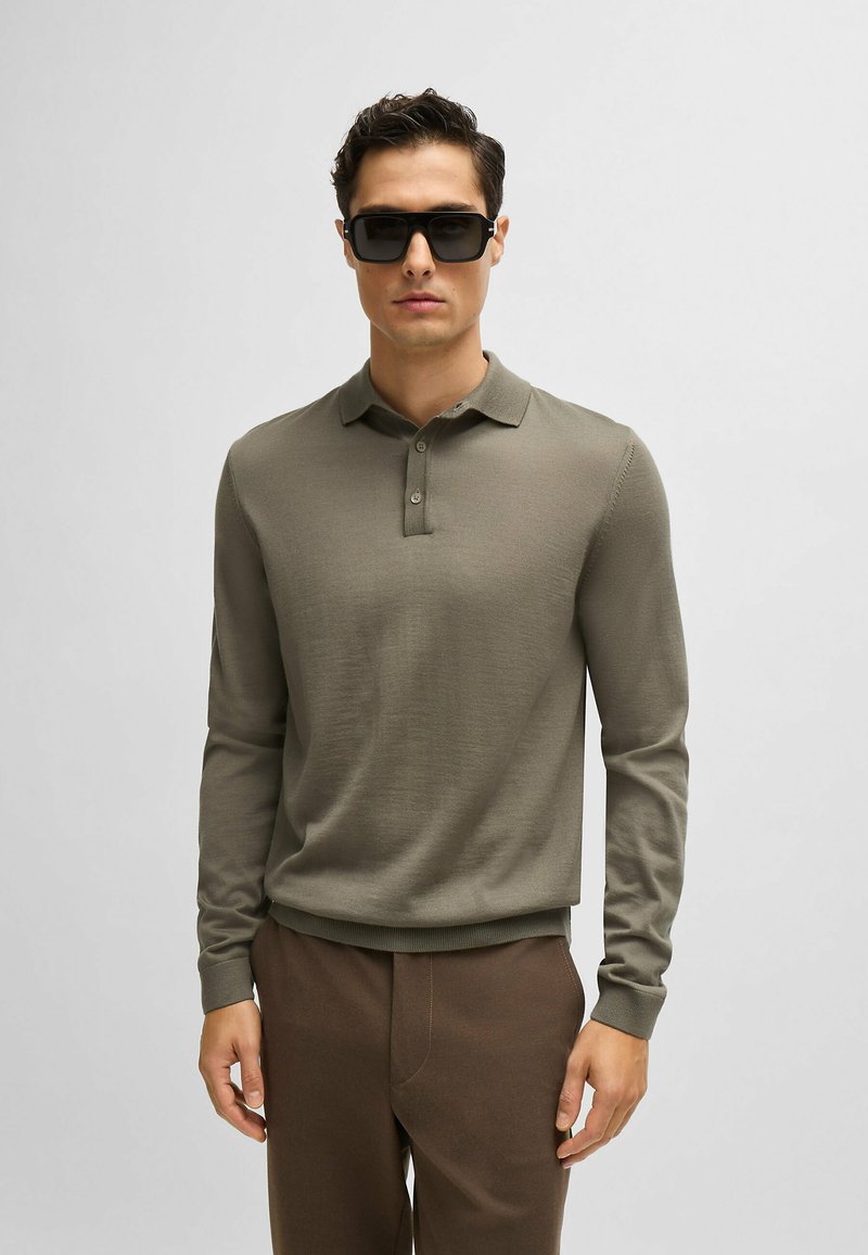 Polo shirt verde oliva a maniche lunghe con texture a costine, colletto con tre bottoni e polsini aderenti, abbinato a pantaloni marroni. Occhiali da sole neri.