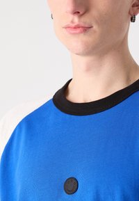 Blue short-sleeve shirt with black collar and white shoulders, featuring a small black logo on the front. Smooth cotton texture.